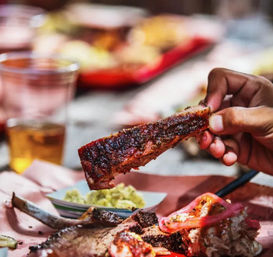 Close-up of a hand holding a smoky, pepper-rubbed barbecue rib over a paper-lined tray with pickled onions and a small side — juicy smoked pork ribs, close-up BBQ shot.