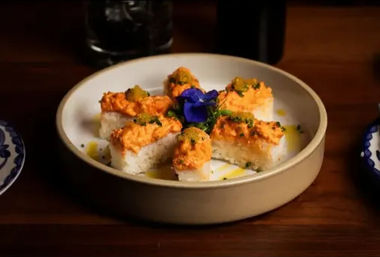 Japanese-inspired sushi appetizer: six rice squares topped with creamy orange seafood mixture, herbs and oil, arranged around a purple edible flower on a beige plate