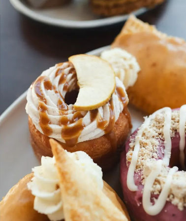 Close-up of assorted gourmet donuts on a plate: caramel-apple cream donut with whipped frosting, caramel drizzle and apple slice; purple berry-glazed donut with crumb topping and white icing; cream-filled pastry piece.
