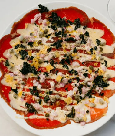 Italian-style beef carpaccio on a white plate — thin red slices drizzled with creamy sauce, scattered capers, diced shallots, chopped herbs and grated egg for a bright, colorful appetizer.