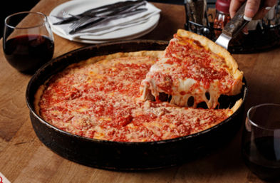 Chicago-style deep-dish cheese pizza in a black pan on a wooden table, a slice lifted showing gooey mozzarella pull, with glasses of red wine and stacked plates nearby