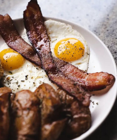 Hearty diner-style breakfast plate with two peppered sunny-side-up eggs, two crispy bacon strips crossed, and browned sausages