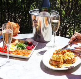 Sunny outdoor patio brunch with two flutes of sparkling wine, chilled bottle in an ice bucket, smoked salmon and bagel platter, and hands cutting eggs Benedict topped with hollandaise