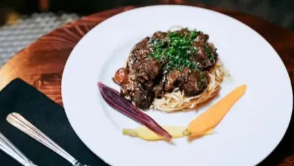 Gourmet braised beef over angel-hair pasta topped with chopped parsley, plated with purple, yellow and orange baby carrots on a white plate at a rustic wooden table