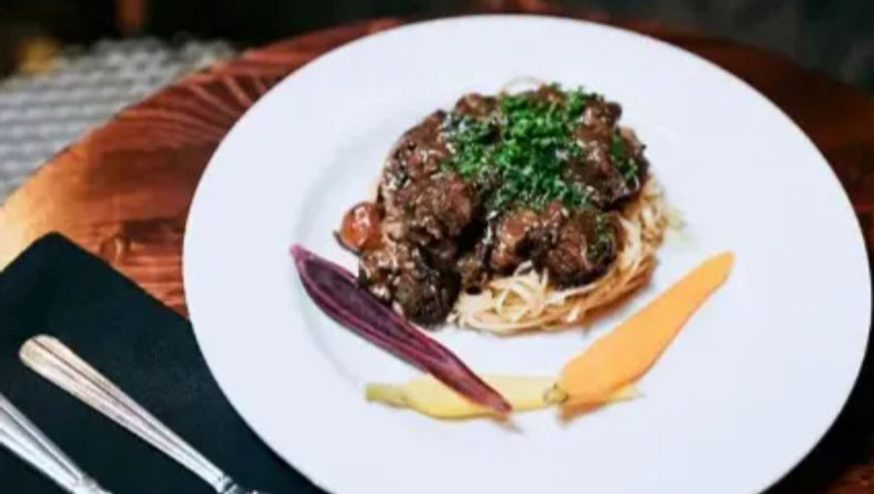 Gourmet braised beef over angel-hair pasta topped with chopped parsley, plated with purple, yellow and orange baby carrots on a white plate at a rustic wooden table