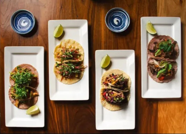 Top-down shot of four white plates of colorful Mexican street-style tacos with shredded meat, slaw, lime wedges and blue dipping bowls on a wooden table