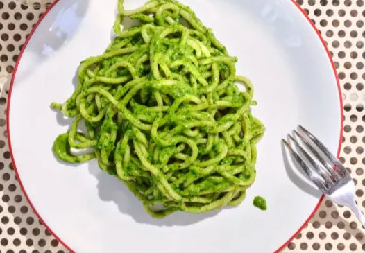 Top-down view of bright green pesto spaghetti on a white plate with a red rim, fork to the side on a perforated tabletop