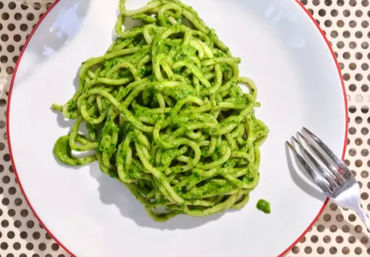 Top-down view of bright green pesto spaghetti on a white plate with a red rim, fork to the side on a perforated tabletop