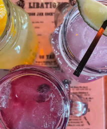 Overhead shot of colorful mason-jar cocktails — pink and purple drinks with ice and lime and lemon garnishes on a pink menu at a cocktail bar.