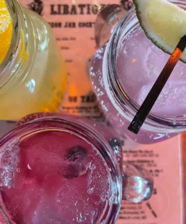 Overhead shot of colorful mason-jar cocktails — pink and purple drinks with ice and lime and lemon garnishes on a pink menu at a cocktail bar.