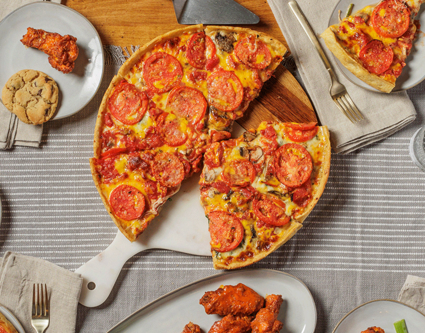 Top-down pepperoni pizza topped with tomato slices on a wooden paddle, surrounded by buffalo chicken wings, a cookie and plates on a striped tabletop — casual dinner spread.