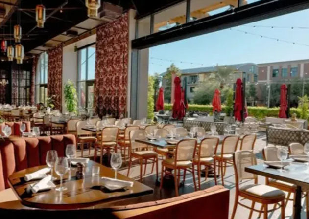 Inviting modern indoor-outdoor restaurant with open wall to a sunny al fresco patio, red umbrellas, neatly set tables with wine glasses, cushioned banquettes and wooden chairs.