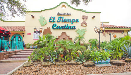 Southwest-style Mexican cantina exterior with colorful facade, turquoise wrought-iron entrance, red tile roof, decorative cactus and hourglass accents, vintage lamp posts and a lush agave and succulent garden in a rocky front bed.