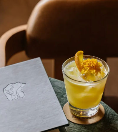 Vibrant yellow citrus cocktail on ice in a rocks glass, garnished with an orange peel and edible yellow flower, sitting on a coaster beside a minimalist menu on a marble table with warm leather seating in the background.