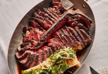 Juicy sliced T-bone steak, grilled with char marks and pink center, served on an oval metal platter with roasted bone marrow topped with pickled onions and herbs on a white tablecloth.