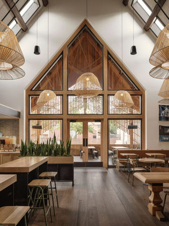 Sunlit modern cafe interior with vaulted A-frame timber windows and glass doors, oversized rattan pendant lights, skylights, wooden floors and tables, green planters and bar stools with a view to an outdoor patio