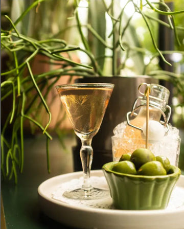 Amber cocktail in a vintage coupe glass beside a green bowl of olives and a small bottle on crushed ice, leafy indoor bar setting