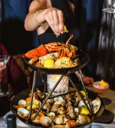 Two-tier seafood tower piled with a whole lobster, clams and lemon wedges as a hand squeezes fresh lemon over it on a candlelit restaurant table.