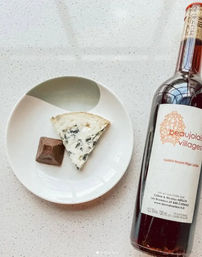 Top-down view of a white plate on a speckled countertop with a wedge of blue cheese and a chocolate square beside a bottle of red wine labeled Beaujolais Villages.