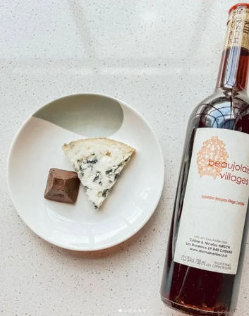 Top-down view of a white plate on a speckled countertop with a wedge of blue cheese and a chocolate square beside a bottle of red wine labeled Beaujolais Villages.