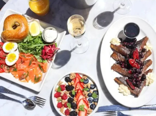 Sunlit brunch spread on a marble table: smoked salmon bagel with soft‑boiled eggs, arugula and pickled onions; yogurt and granola bowl topped with strawberries, blueberries and blackberries; berry‑topped French toast with syrup and a glass of white wine.