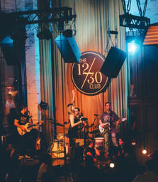 Live band on an intimate indoor stage at a dimly lit music venue — singer with microphone, guitarists and drummer under hanging speakers and warm stage lighting, crowd in foreground.