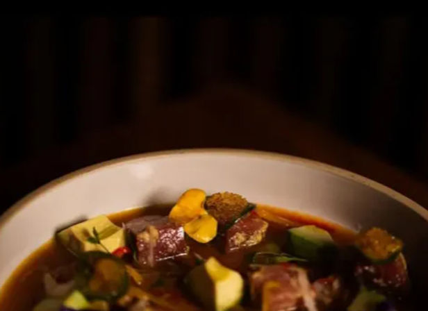 Close-up of a savory poke-style bowl with cubed tuna, avocado, sliced jalapeño, crispy plantain chips and herbs in a rich broth against a cozy dark background