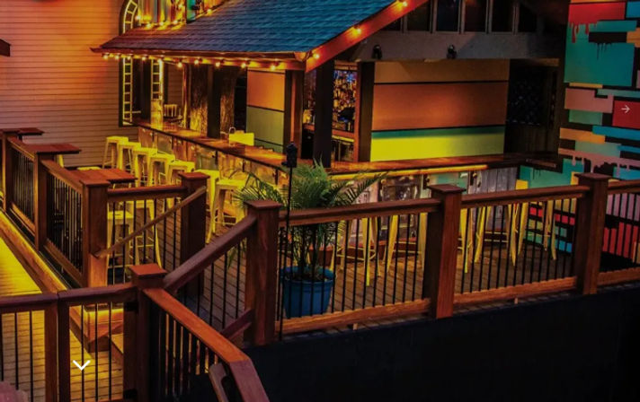 Nighttime outdoor bar on a wooden deck with warm string lights, high bar counter and yellow stools, potted palm, and vibrant painted mural