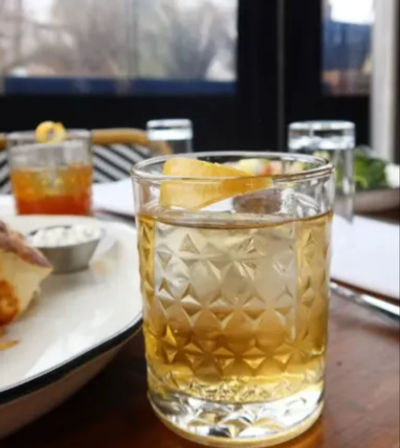 Textured old-fashioned glass holding a golden cocktail with a lemon twist on a restaurant table beside a plate of appetizers and blurred outdoor patio in the background.