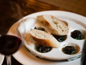 Close-up of a French-style escargot appetizer in a six-well dish with garlic-herb butter, two slices of baguette on top and a blurred glass of red wine
