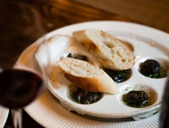 Close-up of a French-style escargot appetizer in a six-well dish with garlic-herb butter, two slices of baguette on top and a blurred glass of red wine