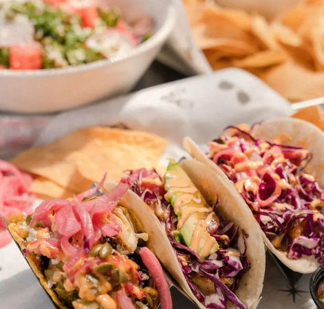Three colorful Mexican-style street tacos in soft tortillas topped with shredded purple cabbage, avocado slices, pickled red onions and spicy crema, served with tortilla chips and a salad bowl in the background