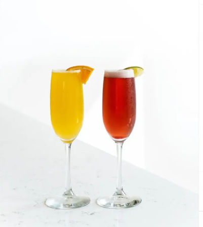 Brunch cocktails: two champagne flutes on a white marble countertop—one bright orange drink garnished with an orange slice and one ruby-red drink topped with foam and a lime wedge against a clean white background.