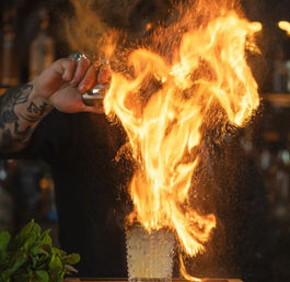 Tattooed bartender igniting a craft flaming cocktail—towering orange flames and sparks erupt from a textured glass on a bar counter next to fresh mint, dramatic mixology moment