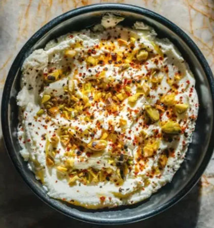Top-down view of creamy whipped yogurt dip (labneh) in a black bowl, drizzled with olive oil and sprinkled with chopped pistachios and red chili flakes on a marble surface.