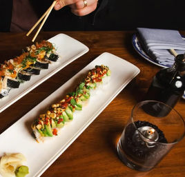 Two long white plates of sushi—avocado-topped roll and seaweed-wrapped maki—on a wooden restaurant table with chopsticks, a soy sauce bottle, folded napkin and a lit candle in a glass.