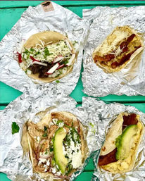 Overhead shot of four assorted Mexican-style tacos wrapped in foil on a turquoise picnic table: beef with radish, onion and cilantro; grilled chicken with avocado and queso fresco; and two breakfast tacos with bacon, egg and shredded cheese.