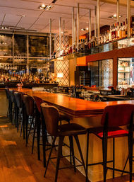 Inviting upscale cocktail bar interior with a long polished wooden counter, leather high stools, hanging glass shelves of liquor bottles, warm ambient lighting and mirrored back wall.