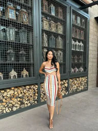 Chic woman in a colorful striped cutout midi dress and heels posing on an outdoor patio by wire‑caged shelves of decorative lanterns with stacked firewood below, holding a tan crossbody bag.