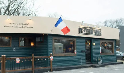 Cozy French bistro exterior with blue-green siding, French tricolor flag, awning noting traditional French cuisine, large windows, string lights on a small wooden patio and adjacent parking lot.