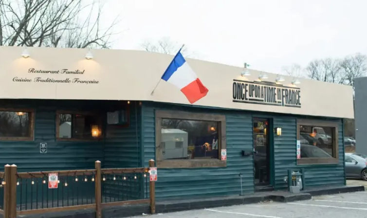 Cozy French bistro exterior with blue-green siding, French tricolor flag, awning noting traditional French cuisine, large windows, string lights on a small wooden patio and adjacent parking lot.