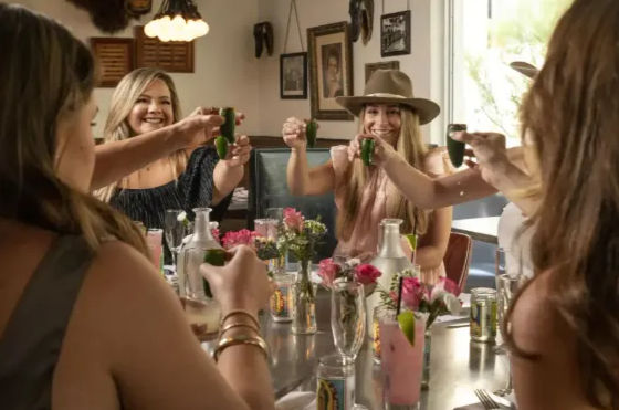 Group of women friends toasting green shot glasses at a sunny restaurant table with pink cocktails, fresh flowers, and one wearing a wide-brim hat