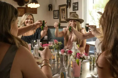 Group of women friends toasting green shot glasses at a sunny restaurant table with pink cocktails, fresh flowers, and one wearing a wide-brim hat