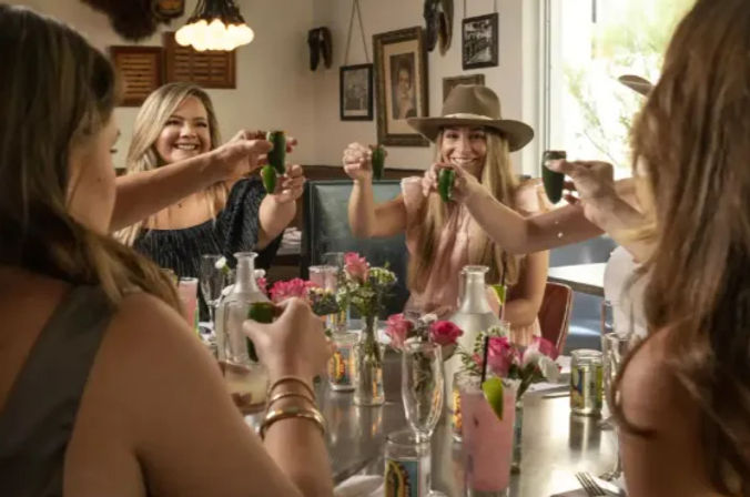 Group of women friends toasting green shot glasses at a sunny restaurant table with pink cocktails, fresh flowers, and one wearing a wide-brim hat