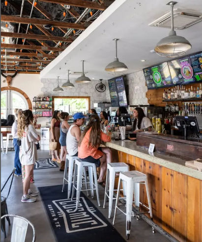 Busy Austin, TX café-bar interior with customers at a wooden counter on white metal stools, industrial pendant lights, exposed ceiling beams and colorful digital menu boards
