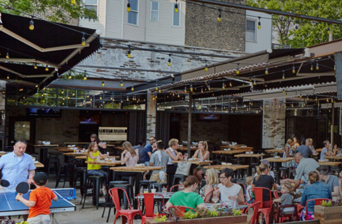 Urban outdoor dining patio with string lights, communal high-top tables and red chairs, families eating and kids playing ping-pong by a brick building