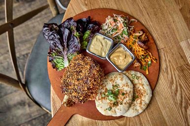 Crispy breaded chicken lollipop on a wooden serving paddle with two grilled flatbreads topped with scallions, lettuce leaves, mixed slaw, shredded carrots and two mustard dipping sauces on a rustic wooden table.