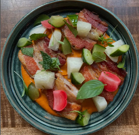 Crispy pork belly slices on roasted red pepper puree with pickled cucumbers, radishes and fresh herbs in a rustic green bowl on a wooden table