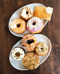 Top-down view of assorted artisan donuts and pastries on two white plates on a wooden table — colorful glazed, frosted, and cream-filled varieties ideal for brunch or cafe menu.