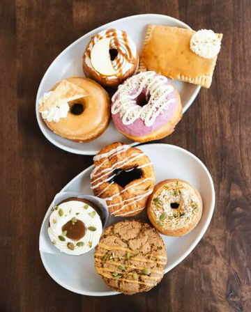 Top-down view of assorted artisan donuts and pastries on two white plates on a wooden table — colorful glazed, frosted, and cream-filled varieties ideal for brunch or cafe menu.